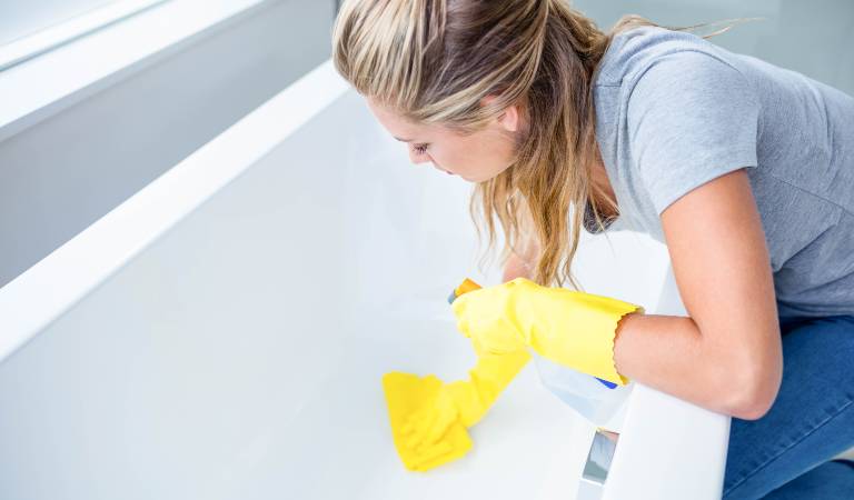 Woman in jeans top and yellow gloves cleaning bathtub using a yellow cloth.