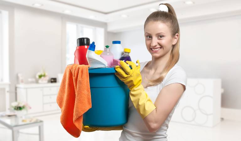 Woman in white top and yellow gloves holding a blue basket filled with tools and supplies.