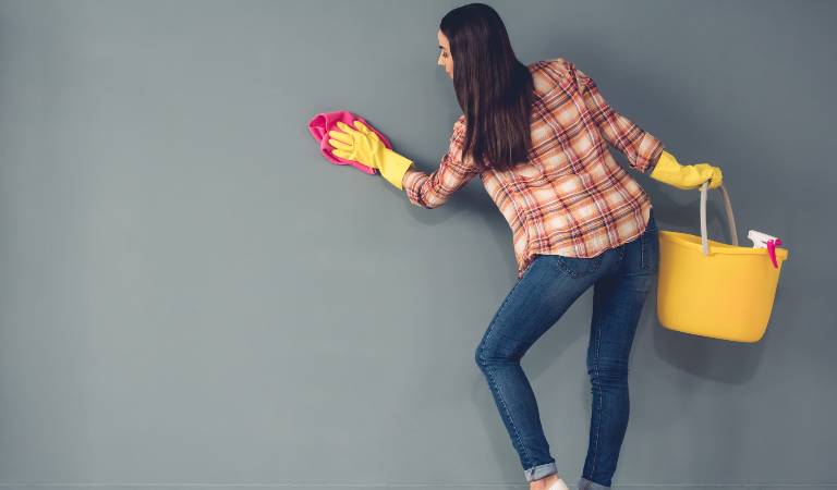 Woman in check shirt, blue jeans and yellow gloves holding a yellow basket and cleaning her wall with red cloth.