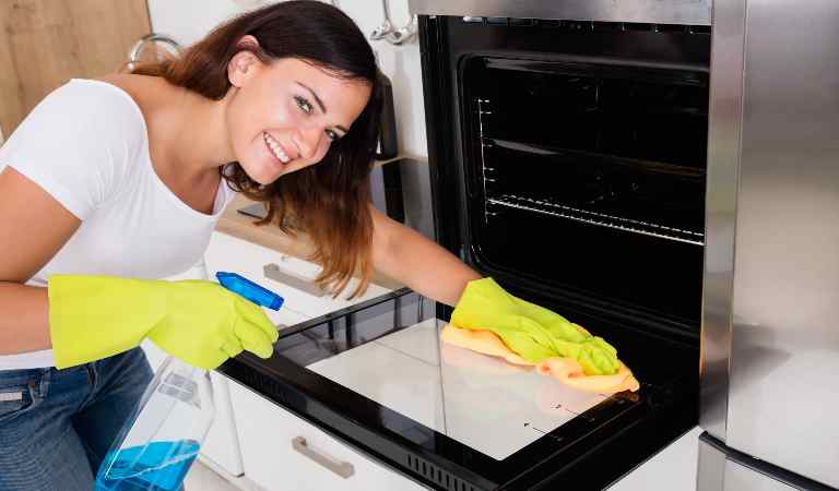 Woman in white top blue jeans and yellow glove cleaning oven inside kitchen