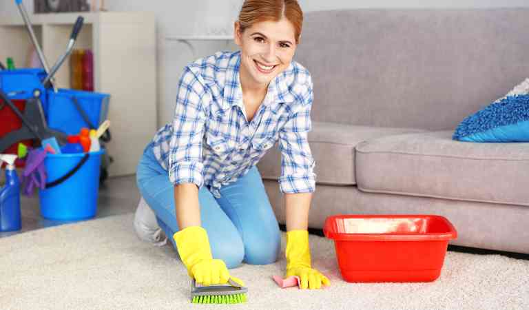 Pretty adult woman in yellow glove cleaning carpet at home using a brush