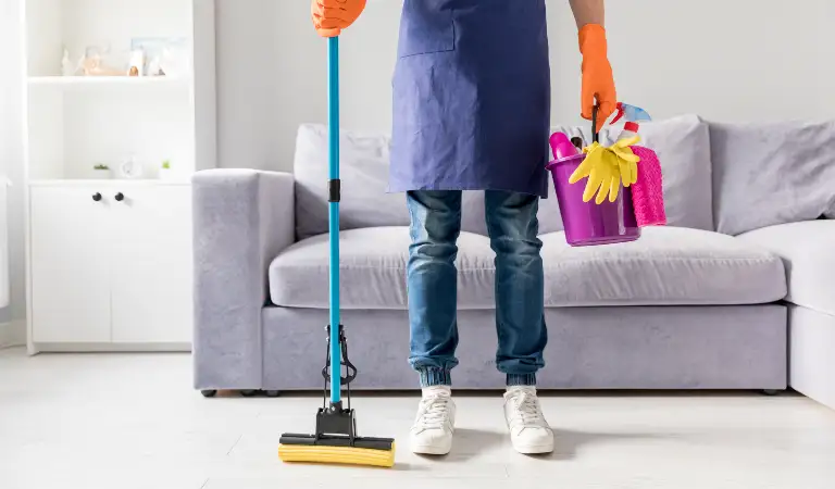 Man in uniform holding a basket in one hand and a mopping stick on his second hand inside his living room.