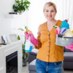 Woman in orange shirt and blue jeans holding a cleaning basket and stick in her hand.