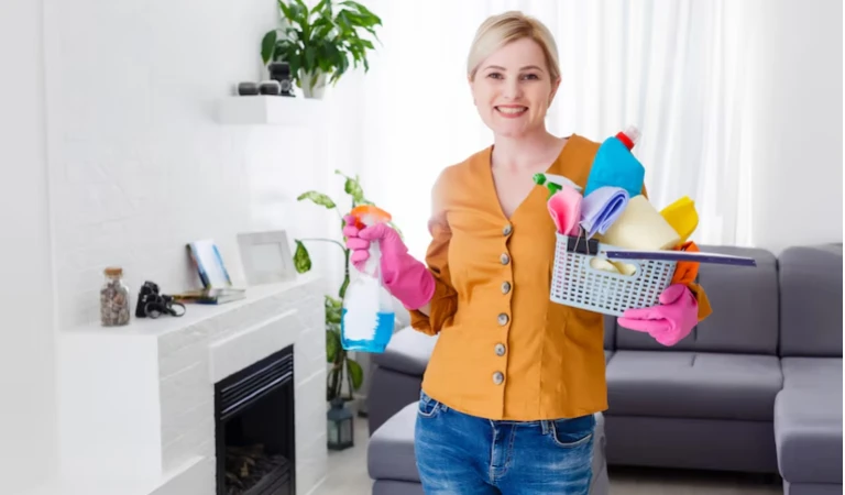 Woman in orange shirt and blue jeans holding a cleaning basket and stick in her hand.