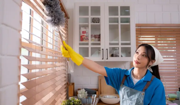 A lady in blue t-shirt and yellow gloves cleaning window blinds using a duster.