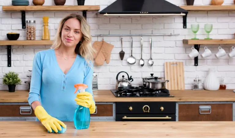 A lady in blue dress and yellow gloves holding a bottle inside her kitchen.