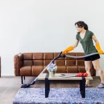 Young woman, housewife in casual clothes and orange rubber gloves using vacuum cleaner to clean floor in living room.