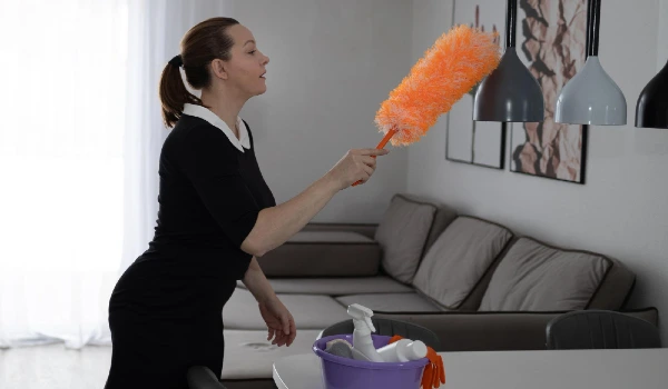 A woman in a uniform cleans a modern house using a feather duster