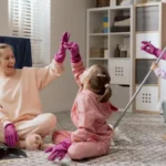 A woman and her two young daughters clean the room mop the floor sweep up the dirt highfive with rubber pink gloves play fool around