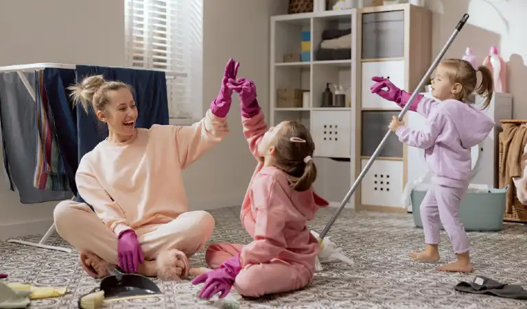 A woman and her two young daughters clean the room mop the floor sweep up the dirt highfive with rubber pink gloves play fool around