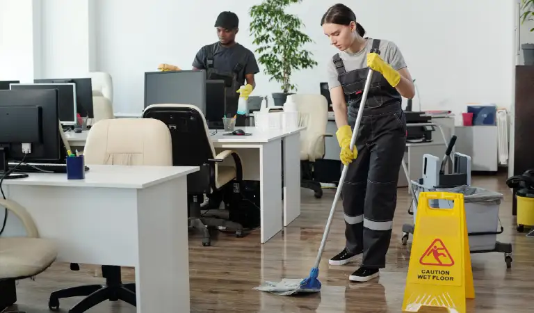 Young black man wiping computer monitors while woman with mop cleaning floor
